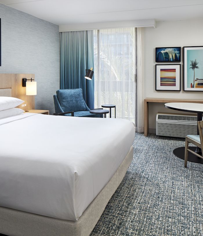 A modern hotel room with a king bed, desk, armchair, blue curtains, framed art, and a table lamp.