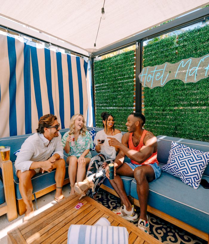 Four friends relax on a hotel patio with drinks, blue-and-white striped curtains, and a green hedge wall reading &ldquo;Hotel Myt&rdquo;.