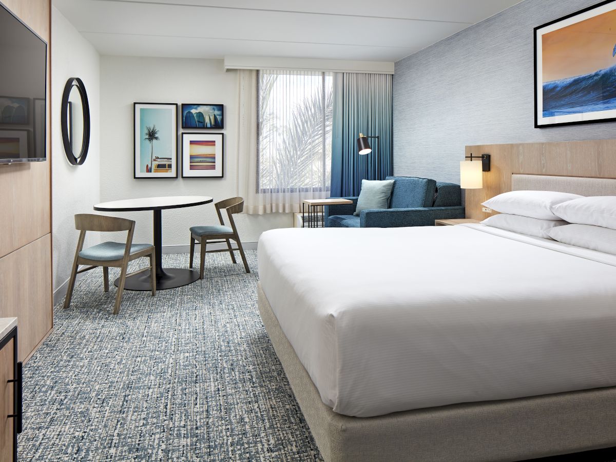 A modern hotel room with a king bed, blue accents, a sitting area by the window, a wall-mounted TV, and framed art; bright and airy.