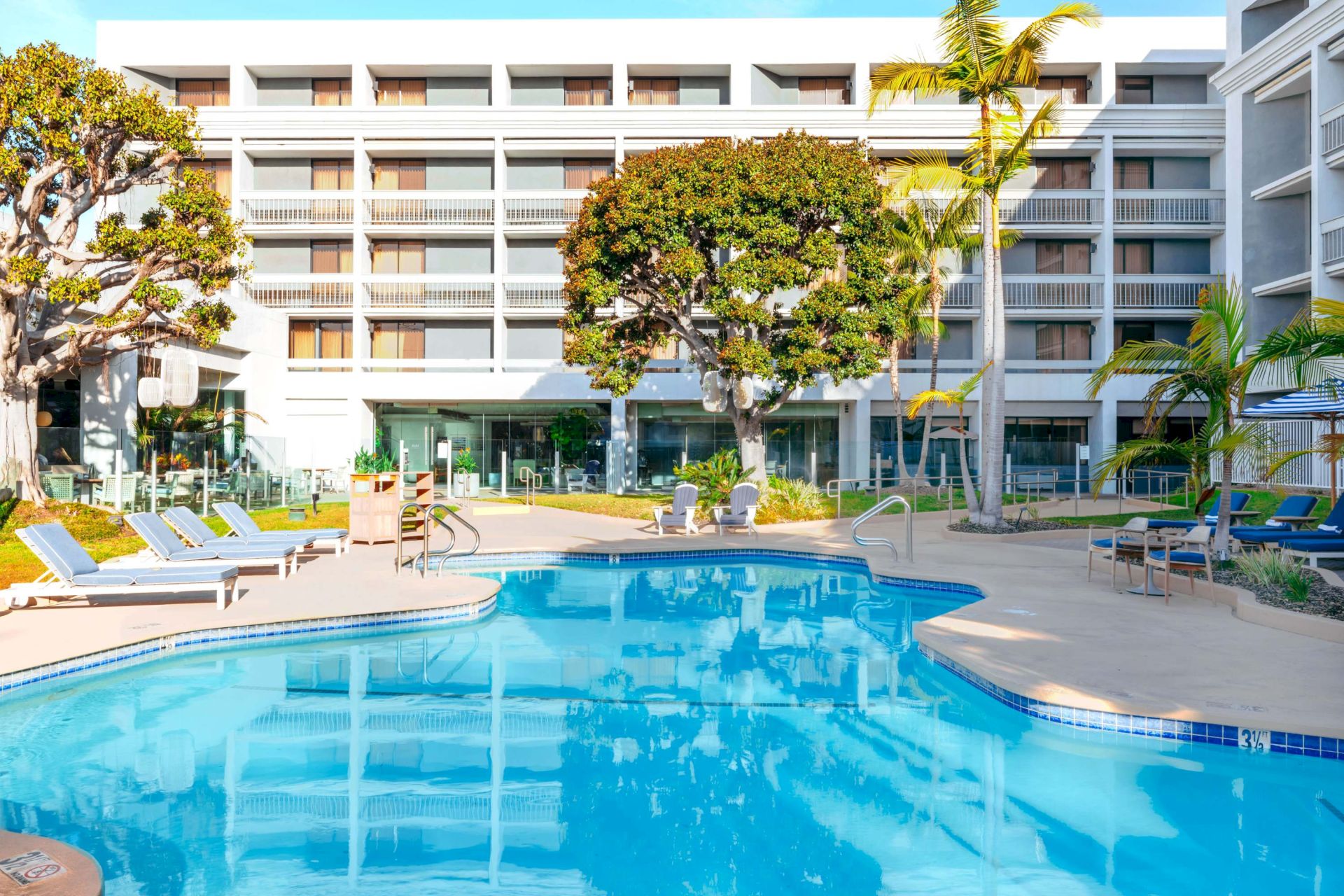 A sunny hotel pool area with a large blue swimming pool, lounge chairs, trees, and a multi-story building in the background.