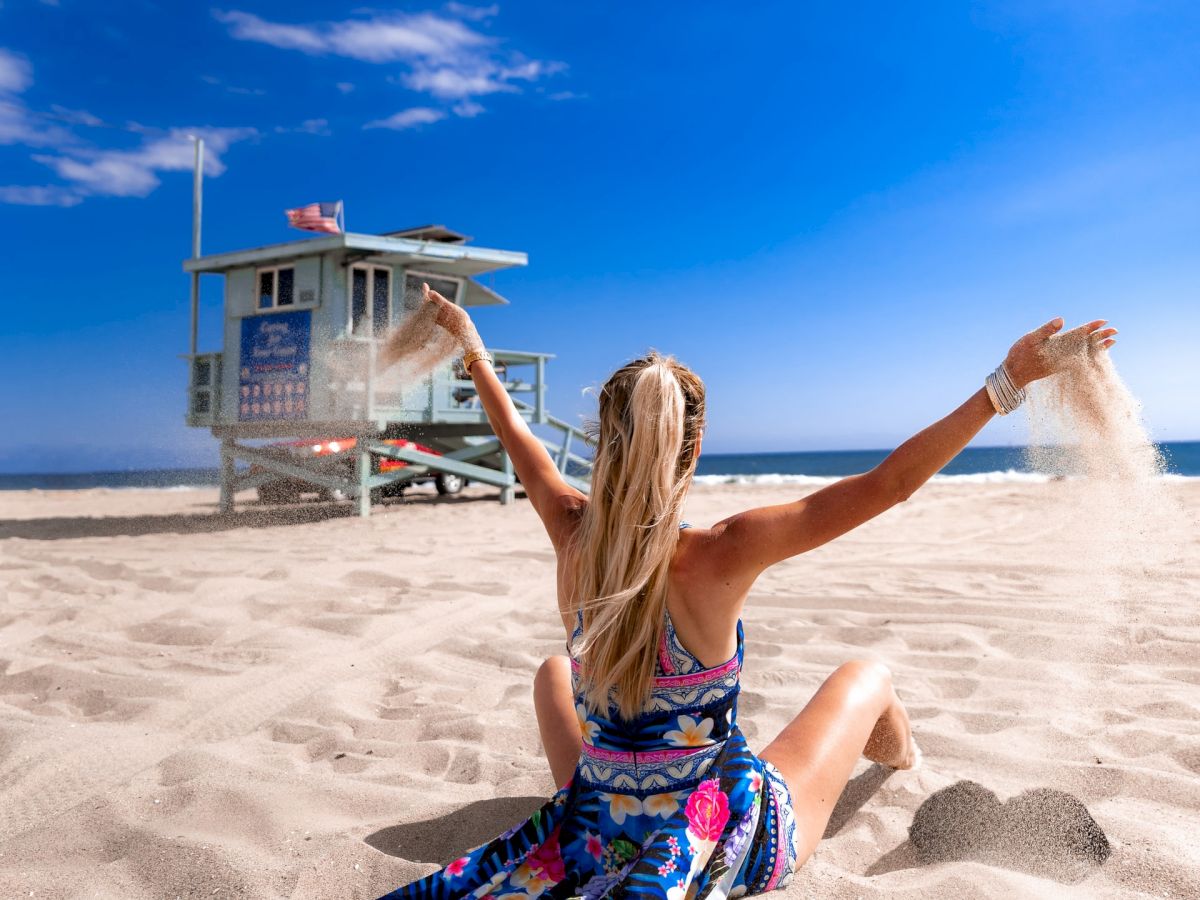 A woman in a colorful dress sits on sunny beach sand, arms raised joyfully toward a lifeguard tower against a bright blue sky.