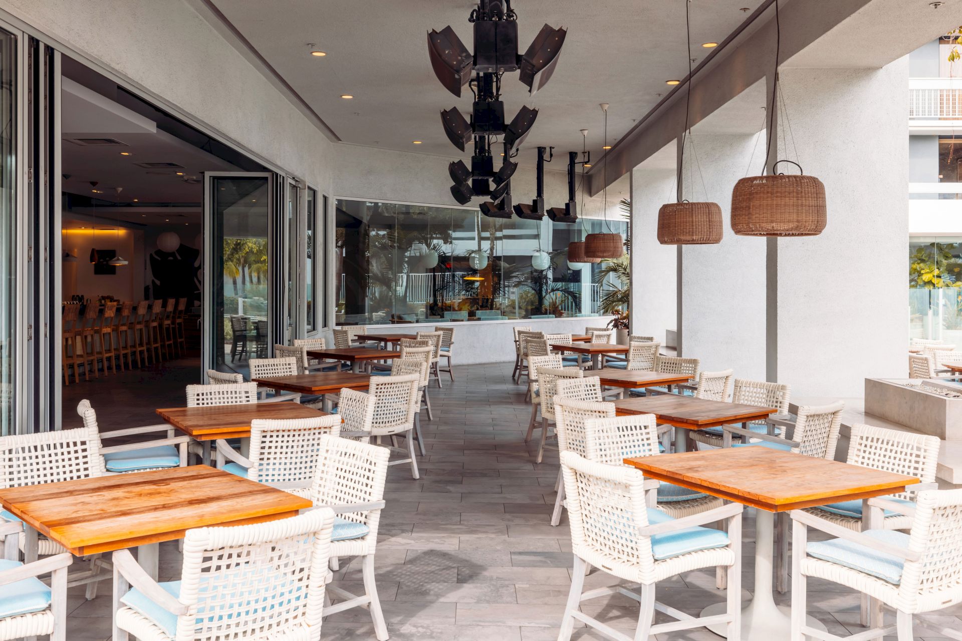 A bright outdoor patio dining area with wooden tables and white chairs, hanging wicker lamps, and a large overhead chandelier.