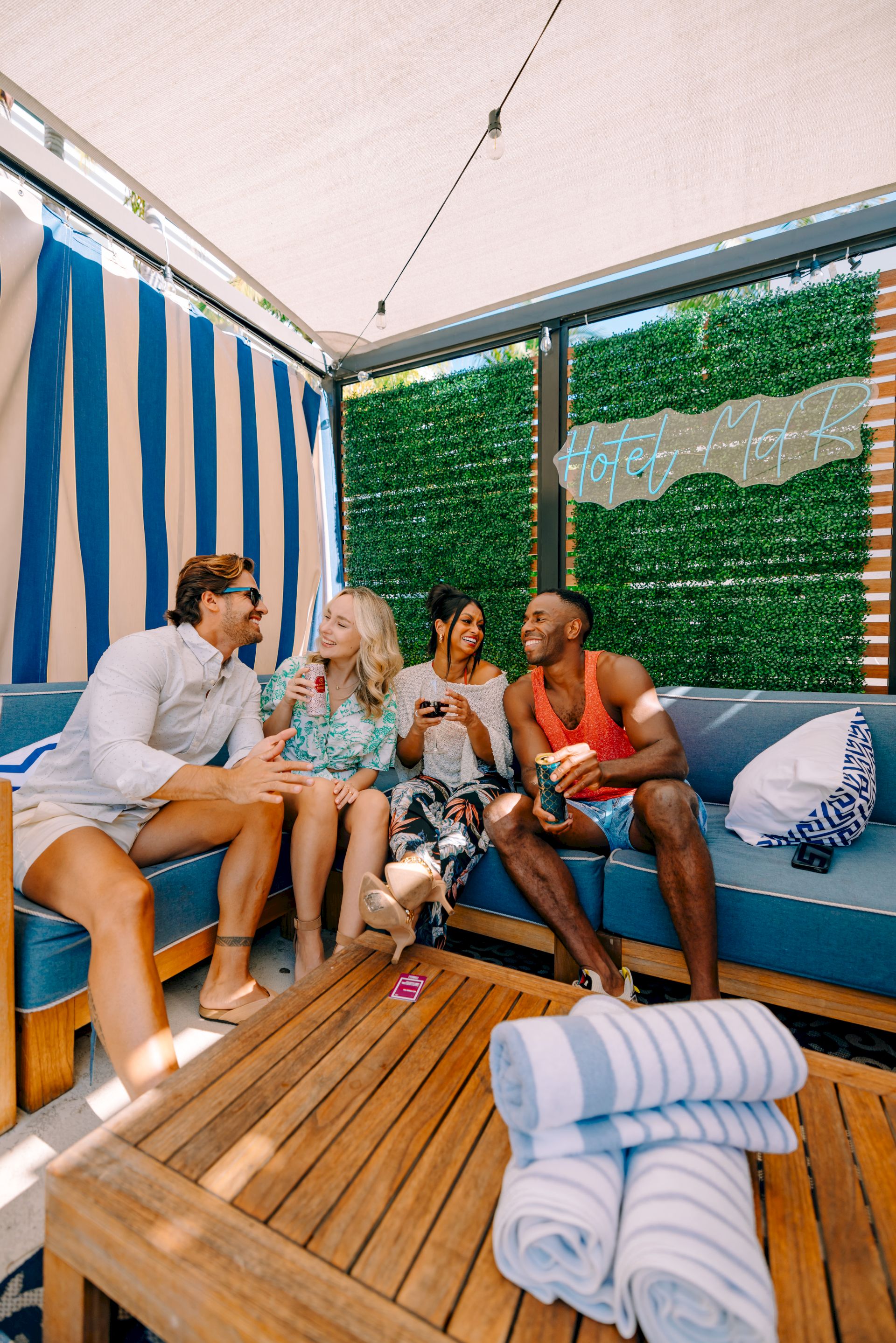 Four friends relax in a hot tub on a sunny patio, laughing with drinks under a green hedge wall reading &ldquo;HotTub&rdquo;.