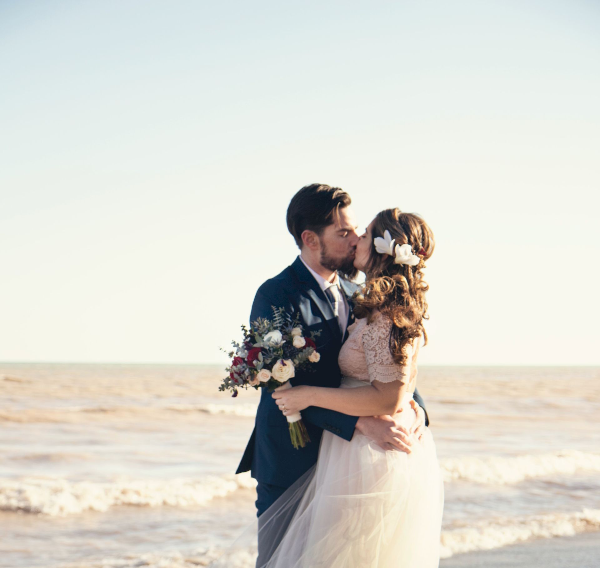 A bride and groom kiss by the seaside, holding a bouquet of flowers as sunlight fills the sky.