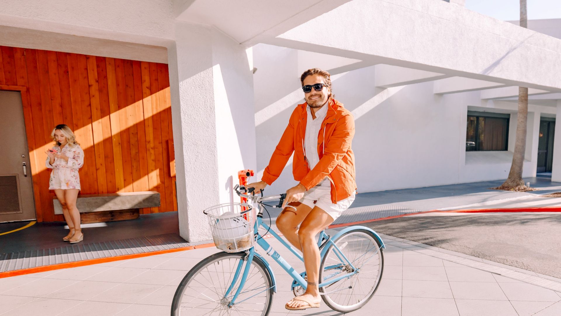 Man in an orange jacket rides a blue bicycle outside a modern building while a woman stands nearby checking her phone.