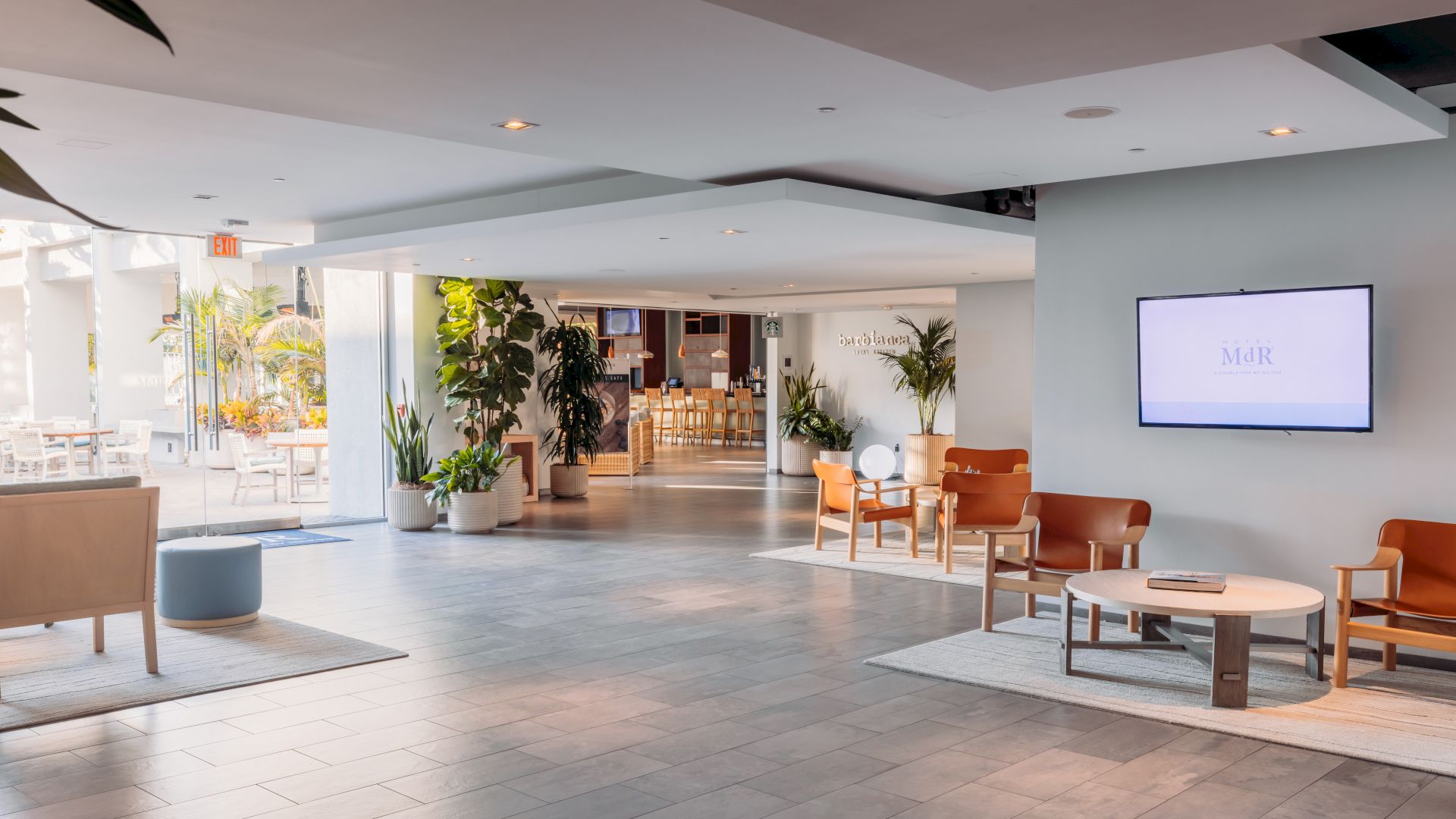 A bright hotel lobby with tiled floors, lounge chairs, potted plants, and a wall TV showing branding and an &ldquo;EXIT&rdquo; sign.