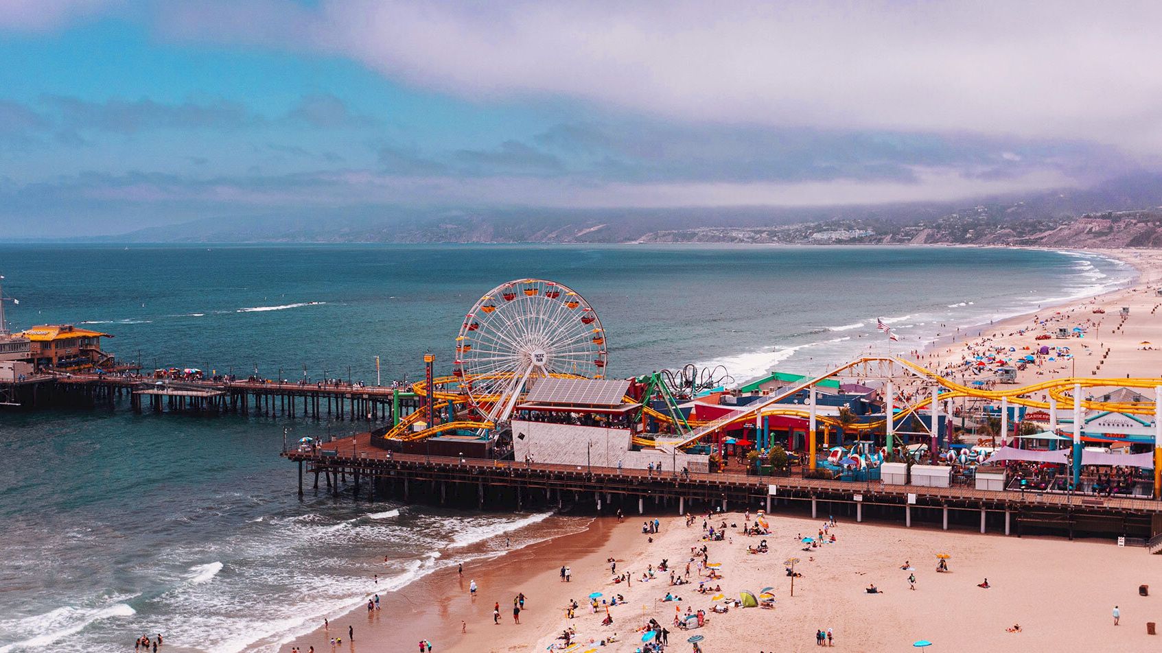 A beach pier with an amusement park, colorful rides, and a giant Ferris wheel beside the ocean at sunset.