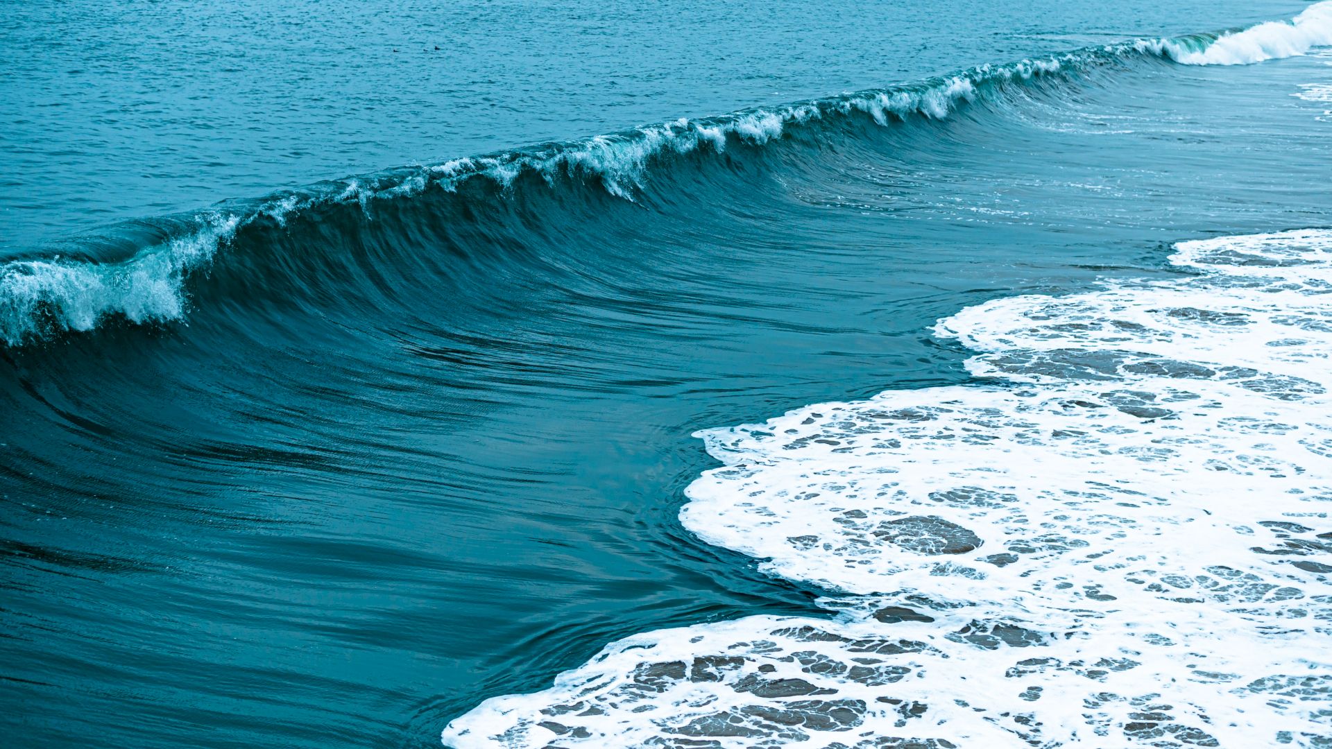 A huge ocean wave crashes onto the shore, sending white foam across the turquoise sea.