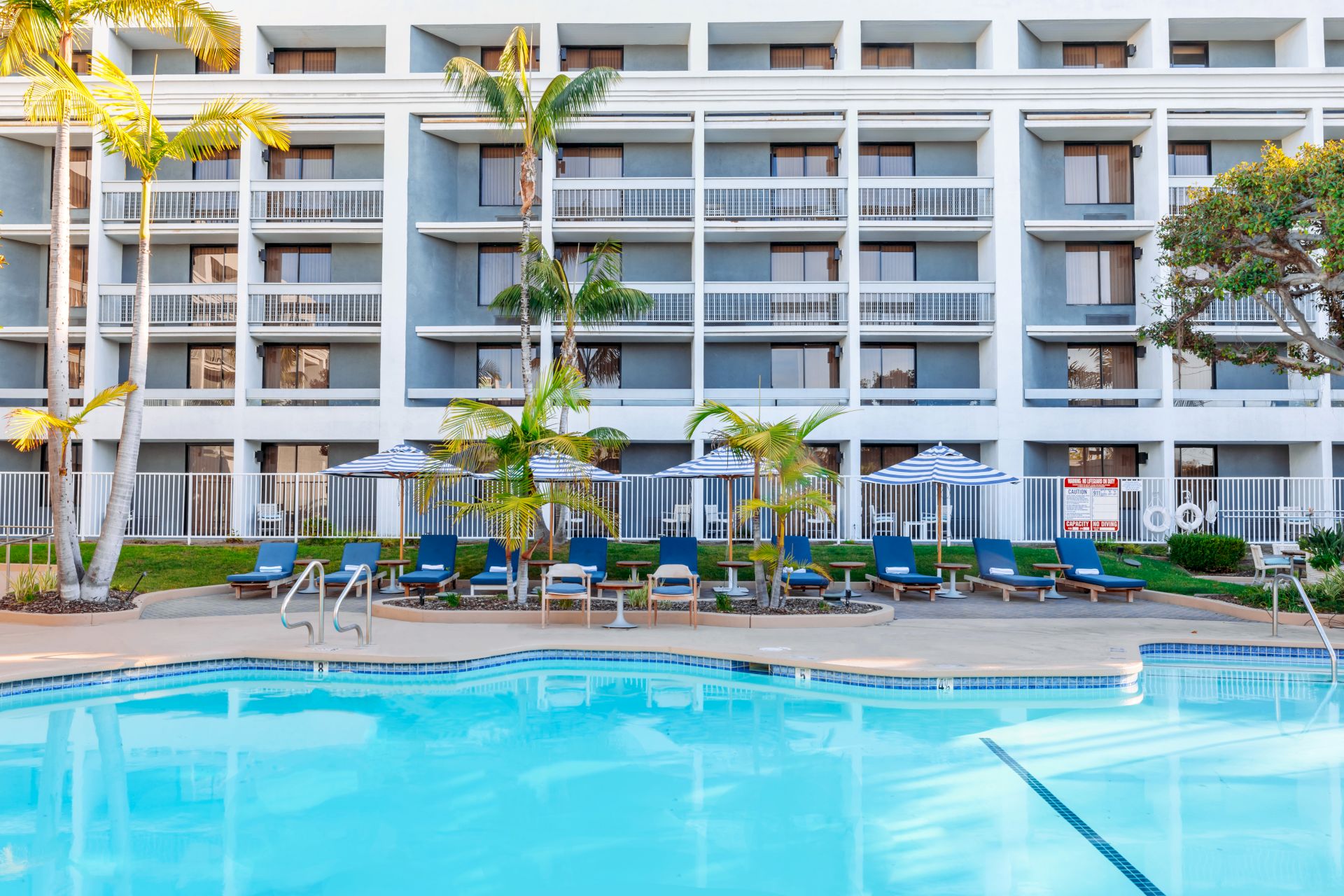 Sunny hotel courtyard with a swimming pool, palm trees, striped umbrellas, and blue lounge chairs in front of apartments.