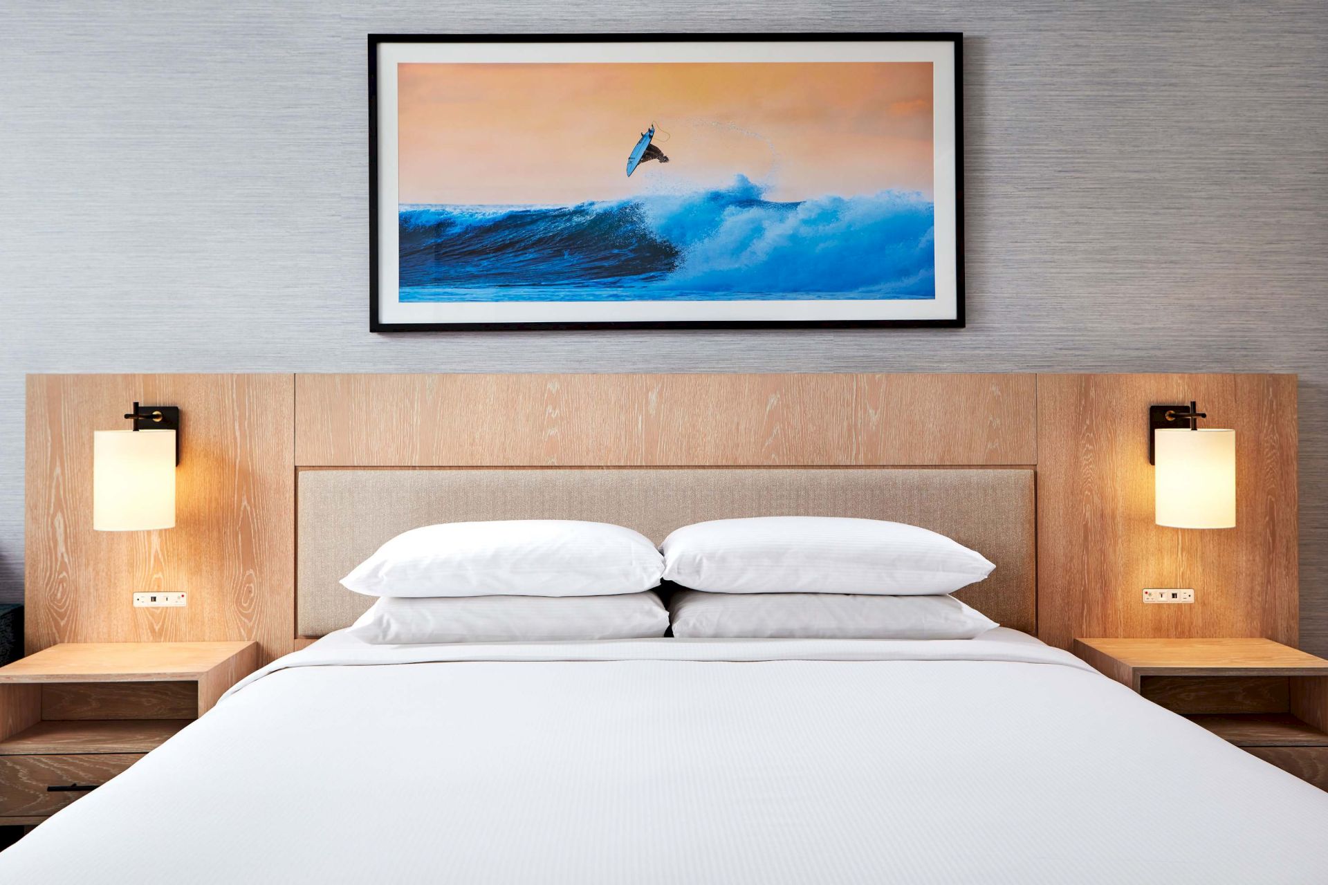 A hotel bedroom with a bed, bedside lamps, wood headboard, and a framed surfing photo above it, adding a coastal vibe.