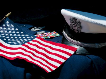 A U.S. flag and a captain&rsquo;s hat on a dark surface, with some small cards or items nearby, suggesting naval or patriotic theme. End sentence.