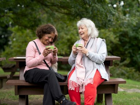 Two elderly women sit on a park bench, sharing smiles and enjoying warm drinks together.
