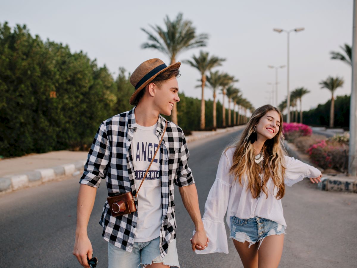 Two friends walk hand-in-hand on a sunny road, smiling and enjoying a casual day out, with palm trees and a clear sky in the background.