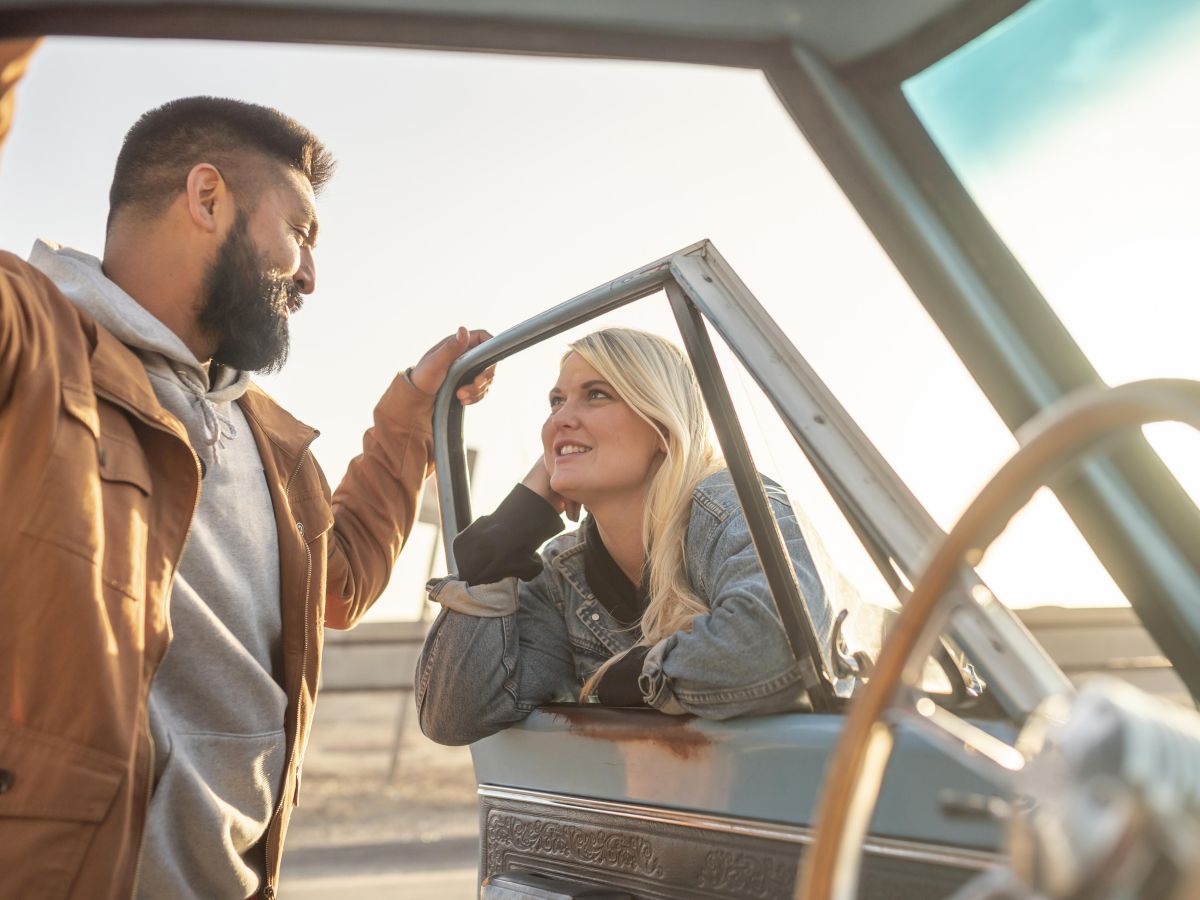 Two people chat beside a vintage car; a man in a brown jacket leans on the door frame while a smiling woman sits inside the truck, posing for a casual moment.