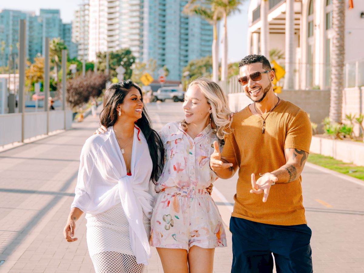 Three friends smile and gesture in a sunny downtown street with tall buildings behind them.